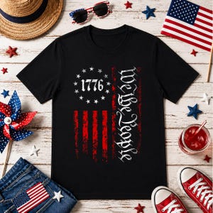 Könnte beinhalten: Schwarzes T-Shirt mit einem verblassten Design der amerikanischen Flagge, dem Text "1776" und "We The People" in Weiß. Das Bild enthält patriotische Dekorationen: eine kleine amerikanische Flagge, Sonnenbrille und rote Turnschuhe.