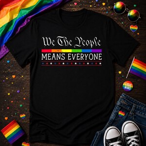 May include: Black t-shirt with the text "We The People MEANS EVERYONE" in white. The text is above a rainbow-colored stripe and a row of stars. The shirt is on a wooden surface with rainbow-themed accessories.