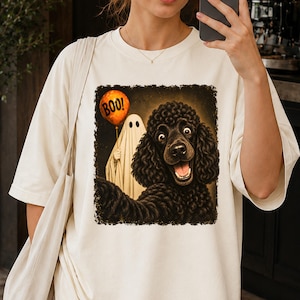 Peut inclure: Un t-shirt couleur crème avec une impression graphique représentant un caniche noir prenant un selfie avec un fantôme tenant un ballon orange. Le ballon porte l'inscription BOO!. Le motif est sur un fond carré sombre et texturé.