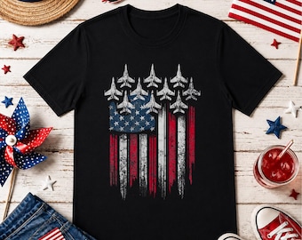 May include: Black t-shirt featuring a distressed American flag design with fighter jets in place of stars. The flag's stripes are red and white. The shirt is surrounded by patriotic accessories, including sunglasses, a flag, and red sneakers.
