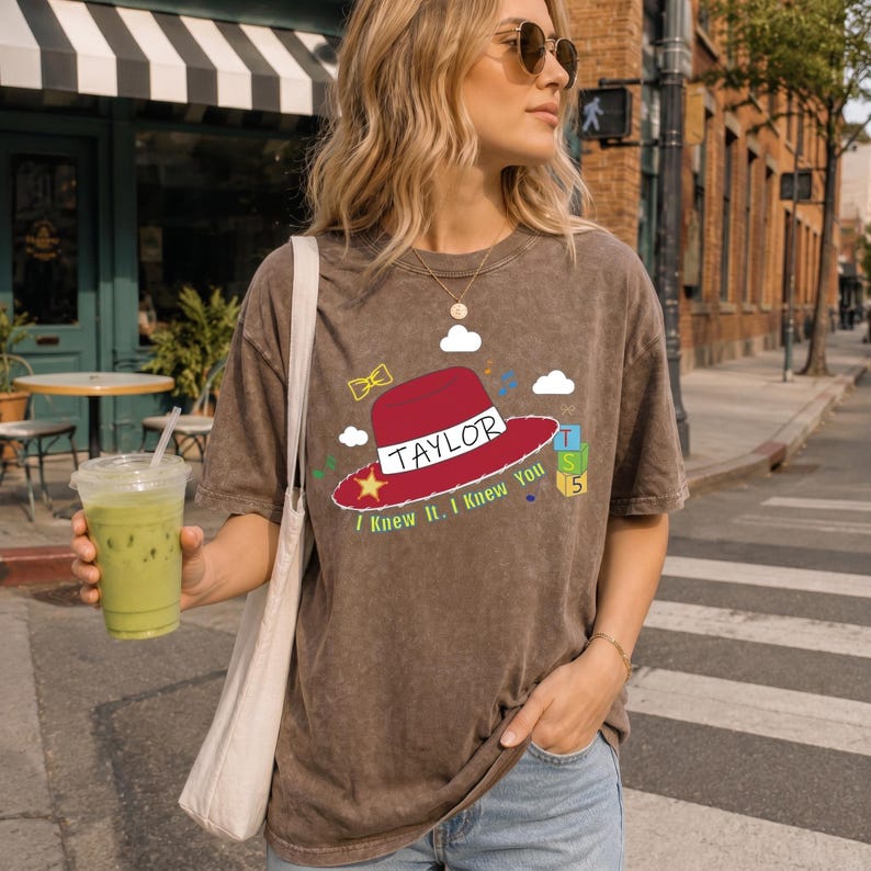 May include: A brown mineral-wash t-shirt featuring a graphic of a red hat with the text TAYLOR and I KNEW IT. I KNEW YOU. The design includes a yellow star, a yellow bow, white clouds, and a stack of three green and blue blocks labeled T, S, 5.
