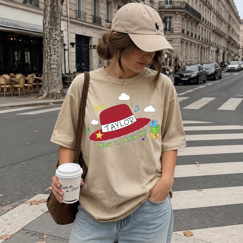 May include: A beige short-sleeve t-shirt featuring a graphic of a red hat with the name TAYLOR printed on it, a yellow star, blue and green blocks with the number 5, white clouds, and musical notes, with the text I KNEW IT, I KNEW YOU below.