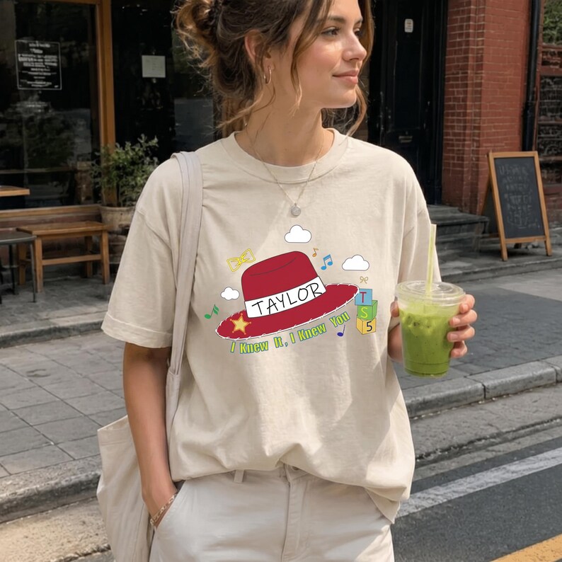 May include: A beige short-sleeved t-shirt featuring a graphic of a red hat with a white band labeled TAYLOR. The design includes the text I Knew It, I Knew You, a yellow star, colorful musical notes, white clouds, and a stack of blocks labeled TS 5.