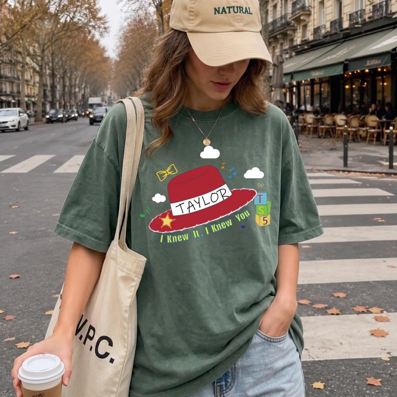 May include: A dark green t-shirt featuring a graphic of a red cowboy hat with the text TAYLOR, a yellow star, a yellow bow tie, musical notes, white clouds, and a stack of blocks labeled T, S, 5, with the text I Knew It. I Knew You below the hat.