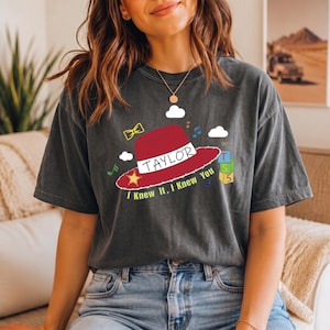 May include: A charcoal gray short-sleeve t-shirt featuring a graphic of a red cowboy hat with a yellow star, the name TAYLOR, and the text I Knew It, I Knew You. The design includes small clouds, musical notes, a bow, and stacked alphabet blocks.