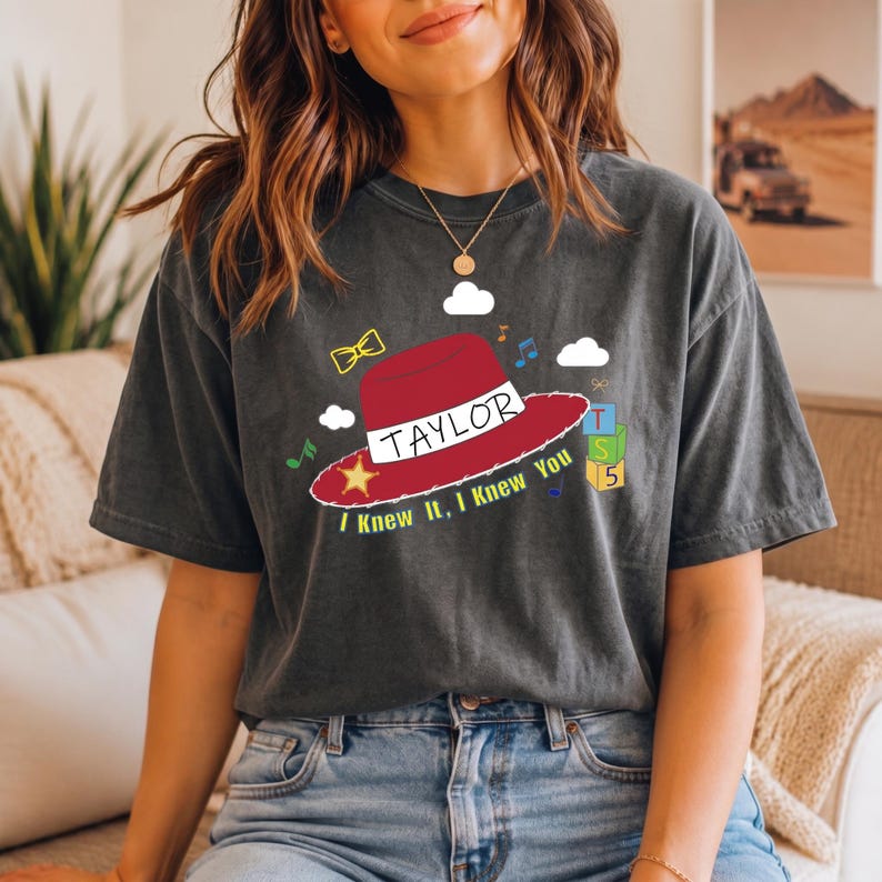 May include: A charcoal gray short-sleeve t-shirt featuring a graphic of a red cowboy hat with a yellow star, the name TAYLOR, and the text I Knew It, I Knew You. The design includes small clouds, musical notes, a bow, and stacked alphabet blocks.