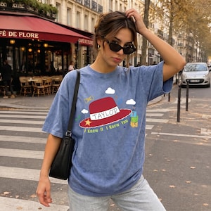 May include: A blue acid-wash t-shirt featuring a graphic of a red hat with the text TAYLOR and I KNEW IT I KNEW YOU, accompanied by small illustrations of clouds, a yellow star, a bow tie, and a stack of green and yellow blocks with the number 5.