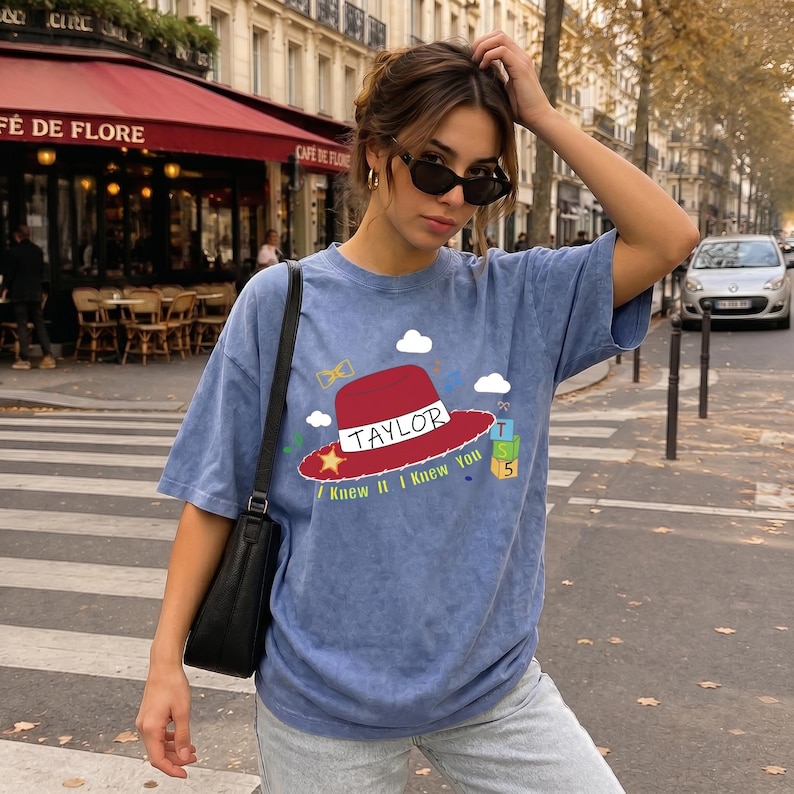 May include: A blue acid-wash t-shirt featuring a graphic of a red hat with the text TAYLOR and I KNEW IT I KNEW YOU, accompanied by small illustrations of clouds, a yellow star, a bow tie, and a stack of green and yellow blocks with the number 5.