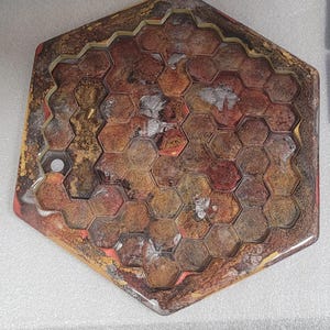 Op de afbeelding: Zeshoekig bordspel met een honingraatontwerp. Het bord heeft een mix van rode, gouden en bruine tinten met een glanzende afwerking. De randen zijn goudkleurig omlijnd. Het bord heeft een heldere, harsachtige coating.