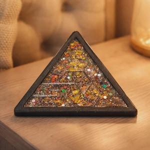 Könnte beinhalten: Ein dreieckiges Dekorationsobjekt mit schwarzem Rand. Das Innere ist mit einem Mosaik aus farbigem Glitzer, Blattgold und kleinen geometrischen Formen gefüllt. Das Objekt liegt auf einer Holzoberfläche.