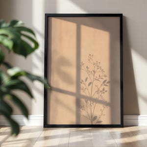 Può includere: Una cornice rettangolare nera contenente una stampa color beige. La stampa presenta la sagoma scura di un delicato stelo di pianta con piccole foglie e boccioli, sovrapposta all'ombra di un telaio di finestra che crea un motivo a griglia.