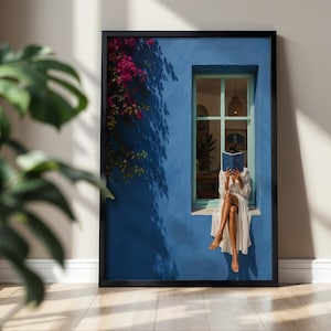 Puede incluir: Una impresión de arte mural enmarcada que muestra una pared azul vibrante con una ventana, una persona con un vestido blanco sentada en el alféizar leyendo un libro azul y flores de buganvilla rosa brillante cayendo por la pared exterior.