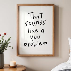 Puede incluir: Un marco de madera rectangular que contiene una impresión blanca con el texto manuscrito en negro That sounds like a you problem. El marco se muestra en una pared, colocado sobre una mesa de madera con un jarrón gris de flores y una vela.