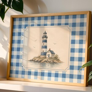 Puede incluir: Una lámina artística enmarcada que presenta una ilustración central de un faro a rayas azules y blancas sobre una isla rocosa, rodeada por un borde de cuadros vichy azules y blancos. La obra está en un marco de madera clara.