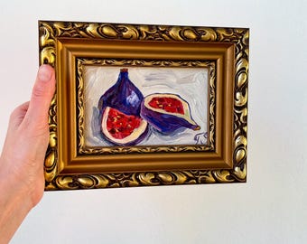 May include: A framed oil painting of figs. The artwork features two whole figs and one sliced open, revealing the red interior. The frame is gold with ornate detailing, and the painting is held by a hand.