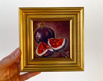 May include: Oil painting of figs in a gold frame. The artwork depicts a whole fig and two fig halves, showcasing the fruit's deep purple skin and vibrant red interior. The frame is a rectangular shape with a gold finish.