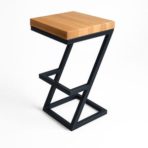 Op de afbeelding: Een moderne barkruk met een licht houten zitting en een zwart metalen Z-vormig frame. De zitting is rechthoekig en lijkt van massief hout te zijn. Het frame is opgebouwd uit hoekige, geometrische vormen.