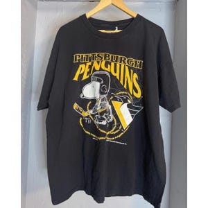 May include: Black t-shirt with a Snoopy character in a Pittsburgh Penguins hockey uniform. The shirt displays the words "Pittsburgh Penguins" in yellow and white, and the team's logo. The t-shirt is hanging on a wooden hanger.
