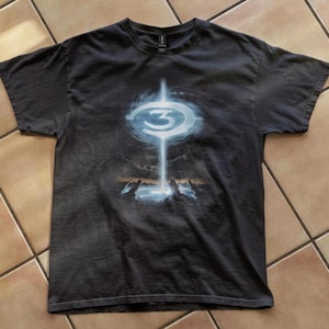 May include: Dark grey t-shirt with a graphic from the video game Halo 3. The design features the number 3 in a blue and white circular emblem, with a vertical light beam and a landscape at the bottom.