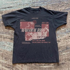 Può includere: Una t-shirt nera a maniche corte con una stampa grafica che riporta il testo MALCOLM TODD, SWEET BOY e CAN'T TAKE YOU HOME YOU HAVE SHOWN THAT YOWON'T GO. Il design include immagini fotografiche di una persona e icone a stella in rosso.