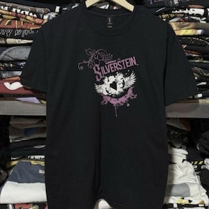 Puede incluir: Camiseta negra de manga corta con el nombre de la banda "Silverstein" en escritura rosa. El diseño incluye un gráfico blanco y rosa con alas y un corazón. La camiseta se muestra en una percha de madera.