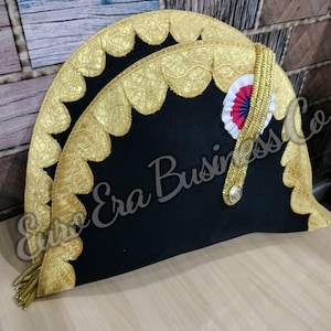 Puede incluir: Un sombrero bicornio negro con ribete dorado y una roseta decorativa. El sombrero presenta un ribete de encaje dorado festoneado a lo largo de los bordes y un detalle de trenza dorada. El texto "Euro Era Business Co" es visible.