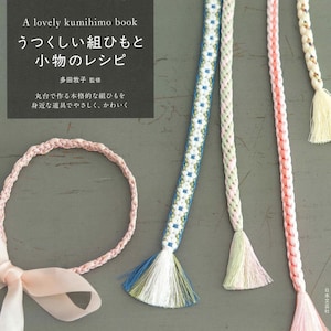 以下が含まれることがあります： 「A lovely kumihimo book」というタイトルの本と、カラフルな組紐。 ピンク、青、緑、白の色合いの紐には、装飾的な房が付いています。 ピンク色の編み込みヘアバンドとリボンも写っています。