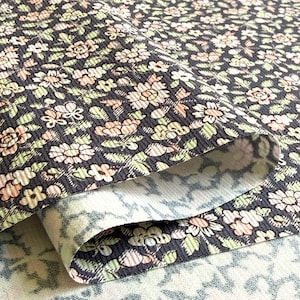 May include: A folded piece of fabric featuring a dark charcoal background with a repeating floral pattern in shades of light pink, cream, and pale green. The material has a distinct ribbed texture across the surface, showing the front and back.