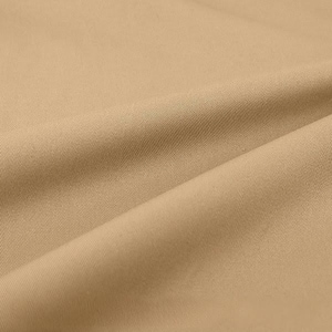 May include: A solid tan fabric with a distinct diagonal twill weave pattern. The material is folded to create soft, flowing curves and shadows, highlighting the uniform texture and consistent light brown colour across the entire surface area.