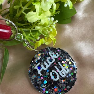 Puede incluir: Un llavero redondo de purpurina negra con las palabras "Fuck Off" en blanco. El llavero tiene un anillo de metal plateado y está rodeado de follaje verde y amarillo.