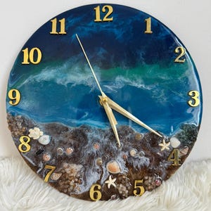 May include: Round wall clock with a beach-themed design. The clock face features a blue and white ocean wave design with gold-colored numerals and clock hands. The bottom of the clock is decorated with seashells and sand.