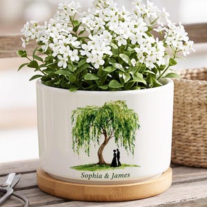 Puede incluir: Una maceta cilíndrica de cerámica blanca con base de bambú, que presenta una ilustración en acuarela de un sauce con una pareja debajo y el texto "Sophia & James" impreso. La maceta contiene una planta con pequeñas flores blancas.