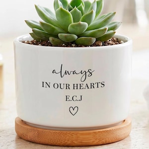 May include: A white ceramic cylindrical planter containing a green succulent plant. The planter features the black printed text "always IN OUR HEARTS" followed by "E.C.J" and a small heart symbol. It rests on a circular light brown bamboo tray.