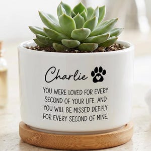 Op de afbeelding: Een witte keramische plantenpot met een bamboe onderzetter en een groene vetplant. De pot is voorzien van de zwarte tekst "Charlie" boven een pootafdruk-icoon en het citaat "YOU WERE LOVED FOR EVERY SECOND OF YOUR LIFE, AND YOU WILL BE MISSED DEEPLY FOR EVERY SECOND OF MINE."