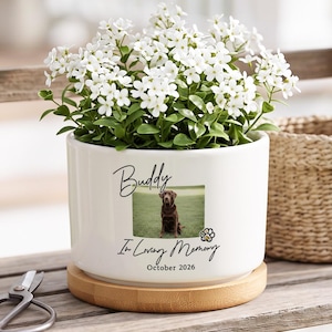 May include: A white ceramic cylindrical planter with a wooden base, containing small white flowers. The planter features a printed photograph of a brown dog, the name Buddy, the text In Loving Memory, the date October 2026, and a paw print icon.