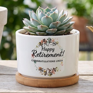 May include: A white ceramic planter with a bamboo saucer containing a green succulent. The planter features a floral wreath graphic with the black text "Happy Retirement!" and "CONGRATULATIONS LINDA" printed on the front surface of the pot.