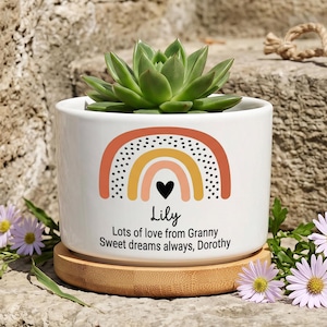 Op de afbeelding: Een witte keramische plantenpot met een bamboe onderzetter en een groene vetplant. De pot is bedrukt met een regenboogafbeelding, een zwart hartje en de tekst: Lily, Lots of love from Granny, Sweet dreams always, Dorothy.