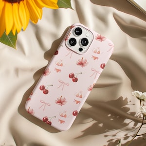 May include: A pale pink phone case adorned with a pattern of red cherries, pink bows, bikini tops and bottoms, and hibiscus flowers. The case has a camera cutout. A yellow sunflower and white flowers are in the background.