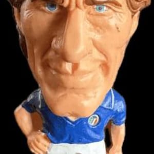 Può includere: Statuetta di un calciatore con una testa grande e tratti esagerati. La figura indossa una maglia blu con colletto e pantaloncini bianchi e scarpe nere. Il giocatore ha i capelli castani e gli occhi azzurri, con un'espressione severa.