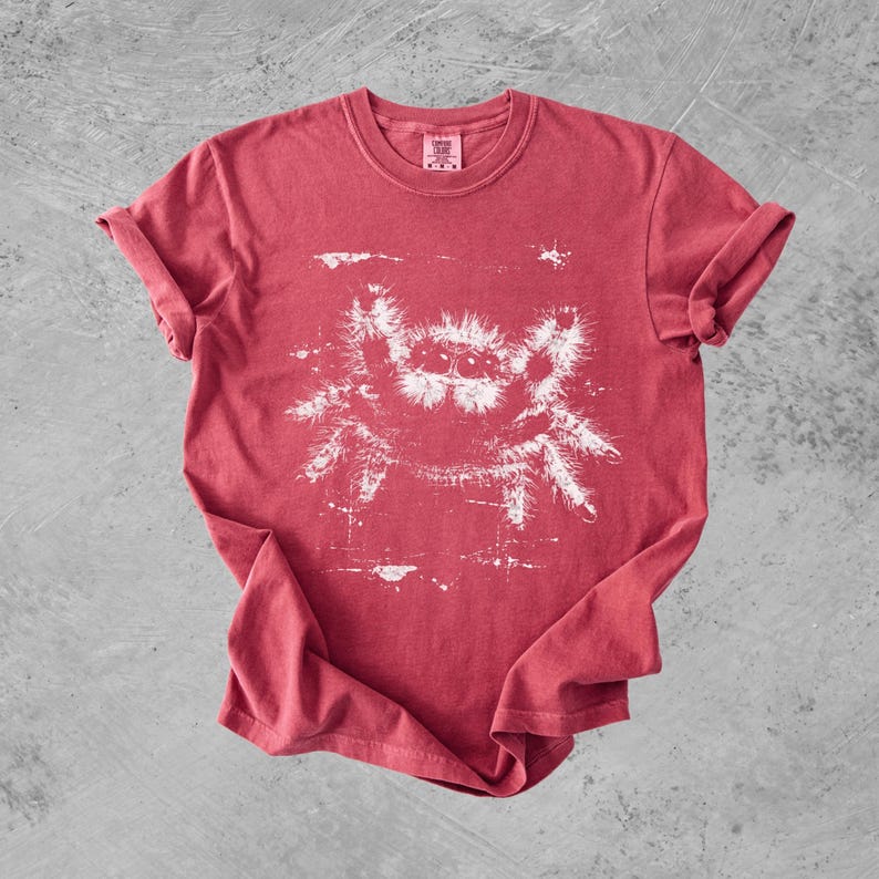 Puede incluir: Una camiseta roja de manga corta con un estampado gr&aacute;fico blanco de una ara&ntilde;a saltarina peluda con ojos grandes. El dise&ntilde;o tiene una textura desgastada y envejecida. La camiseta tiene cuello redondo y una etiqueta en el interior del cuello.