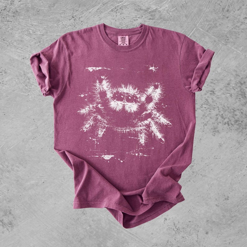 Puede incluir: Una camiseta de manga corta color granate con un estampado gr&aacute;fico blanco de una ara&ntilde;a saltarina peluda en el centro. La camiseta tiene cuello redondo y una etiqueta de la marca Comfort Colors impresa en el interior de la zona del cuello.