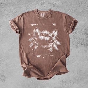 Puede incluir: Una camiseta marr&oacute;n de manga corta con un estampado gr&aacute;fico blanco de una ara&ntilde;a saltarina peluda en el centro. El dise&ntilde;o tiene un aspecto desgastado y envejecido con salpicaduras de pintura blanca alrededor de la ilustraci&oacute;n en el pecho.