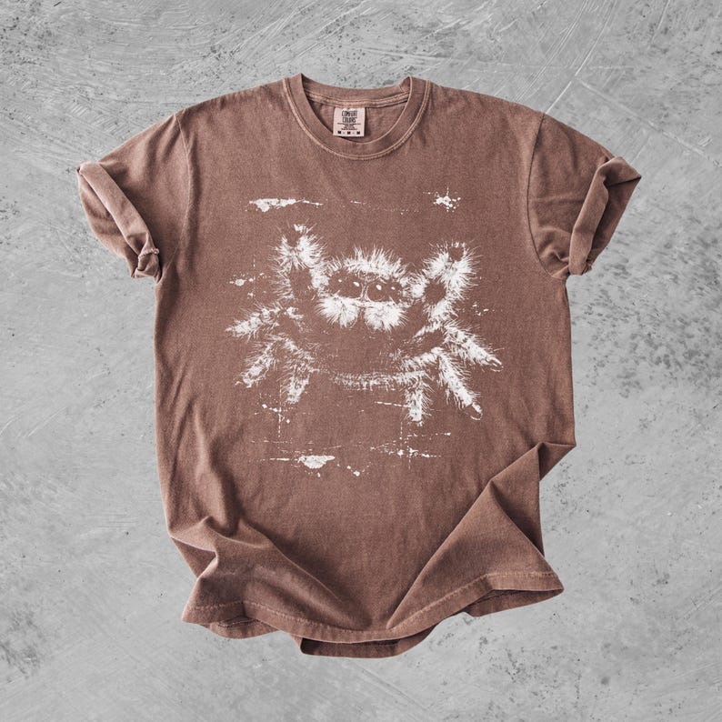 Puede incluir: Una camiseta marr&oacute;n de manga corta con un estampado gr&aacute;fico blanco de una ara&ntilde;a saltarina peluda en el centro. El dise&ntilde;o tiene un aspecto desgastado y envejecido con salpicaduras de pintura blanca alrededor de la ilustraci&oacute;n en el pecho.