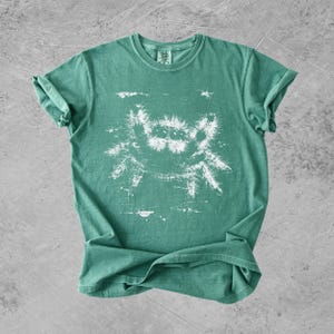 Puede incluir: Una camiseta de manga corta y cuello redondo en color verde espuma de mar apagado. El centro de la prenda presenta una ilustraci&oacute;n gr&aacute;fica blanca con estilo desgastado de una ara&ntilde;a saltarina peluda con varios ojos.