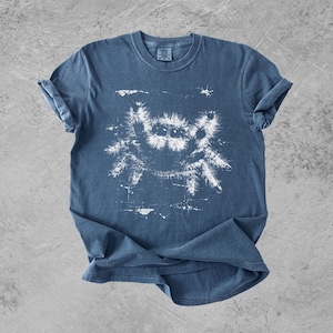Puede incluir: Una camiseta azul de manga corta con un estampado gr&aacute;fico blanco de una ara&ntilde;a saltarina peluda en el centro. La ilustraci&oacute;n de la ara&ntilde;a tiene un aspecto desgastado y texturizado con salpicaduras de tinta visibles alrededor del dise&ntilde;o.