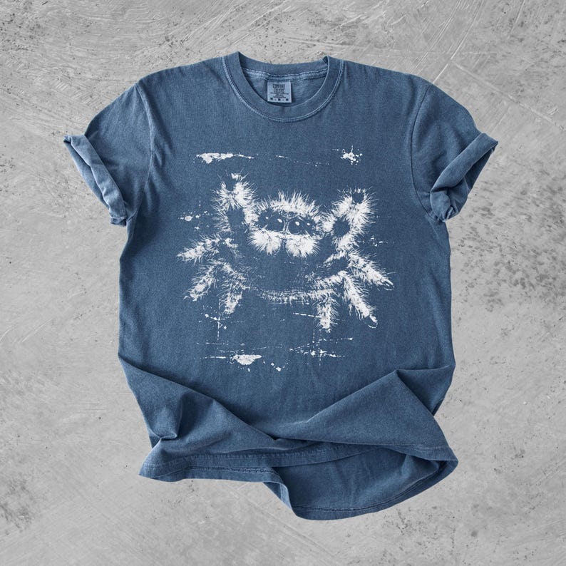 Puede incluir: Una camiseta azul de manga corta con un estampado gr&aacute;fico blanco de una ara&ntilde;a saltarina peluda en el centro. La ilustraci&oacute;n de la ara&ntilde;a tiene un aspecto desgastado y texturizado con salpicaduras de tinta visibles alrededor del dise&ntilde;o.