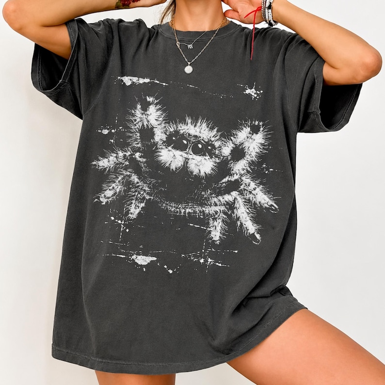 Puede incluir: Una camiseta de manga corta gris carb&oacute;n con un gran estampado gr&aacute;fico blanco desgastado de una ara&ntilde;a saltarina peluda. El dise&ntilde;o de la ara&ntilde;a presenta pelaje texturizado detallado y m&uacute;ltiples ojos visibles en la parte frontal.