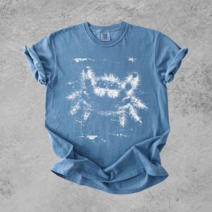 Puede incluir: Una camiseta azul de manga corta con un estampado gr&aacute;fico blanco de una ara&ntilde;a saltarina peluda con ojos grandes. La ilustraci&oacute;n de la ara&ntilde;a tiene un aspecto desgastado y texturizado, y la camiseta tiene cuello redondo y mangas remangadas.