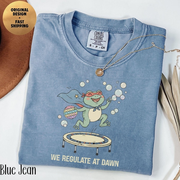 May include: A blue crew neck t-shirt featuring a graphic of a green frog wearing a red superhero cape and mask, jumping on a trampoline while holding a colorful pop-it toy and blowing bubbles. The text "WE REGULATE AT DAWN" is printed below the frog.
