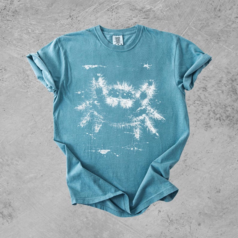 Puede incluir: Una camiseta de manga corta de color azul verdoso con un estampado gr&aacute;fico blanco de una ara&ntilde;a saltarina peluda en el centro. La prenda tiene cuello redondo y un aspecto desgastado y texturizado tanto en la tela como en la tinta blanca.
